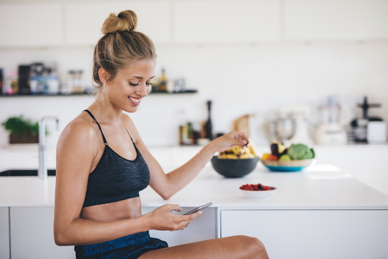 Smiling young woman sitting in kitchen using mobile phone and eating fruits. Female reading text message on her smartphone in morning.