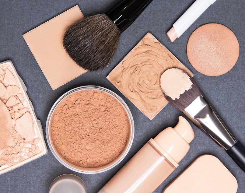 Close-up of concealer pencil corrector open liquid foundation bottle and jar of loose powder crushed compact powder makeup brushes and cosmetic sponges on gray textured surface