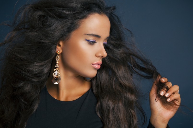 Beautiful model brunette with long curled hair. Profile side. Purple eyeshadow. Pink Lipstick and jewelry earrings. Studio shot over blue background
