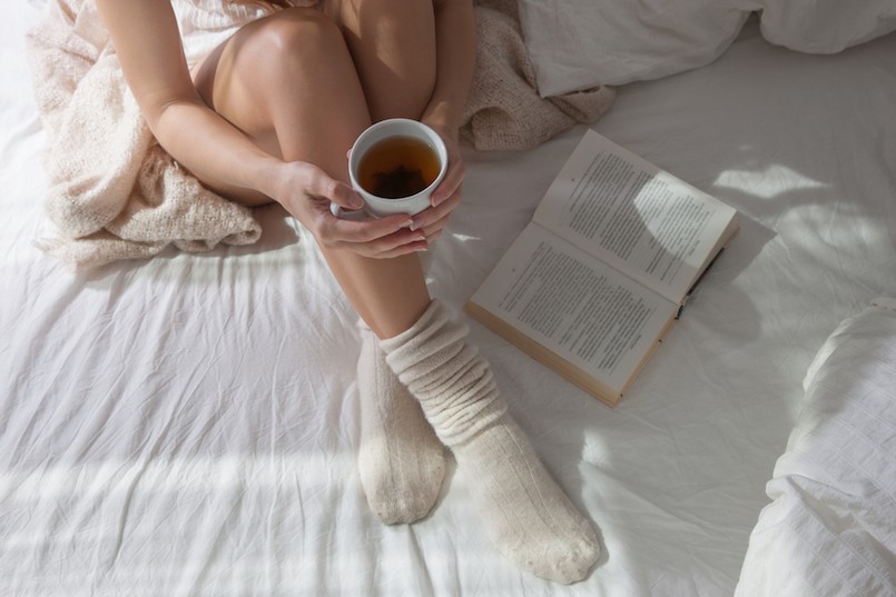 Shapely legs sitting on a bed with a light bed in a warm and cozy bed. A woman sitting on a bed in her hands a cup of hot tea, she reads a book. long female legs in socks