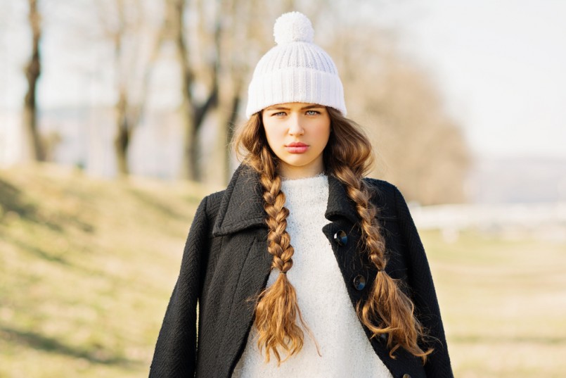 Portrait of gorgeous teenage girl with long blonde braided hair in winter outfit. Beautiful young woman with braids wearing black coat, white sweater and knitted white hat