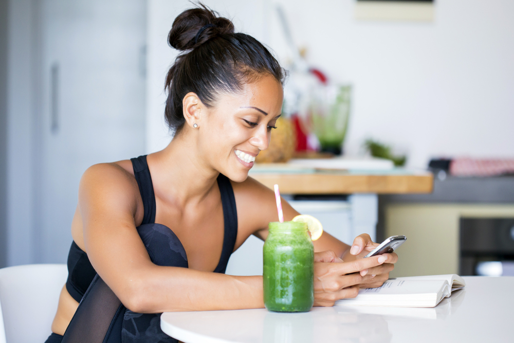 Woman drinking a homemade green detox juice, wearing sportive clothing, texting on her phone while sitting in her kitchen table