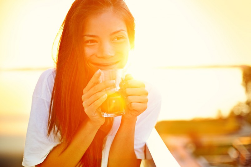 Asian woman drinking coffee in sun sitting outdoor in sunshine light enjoying her morning coffee. Smiling happy multiracial female Asian Chinese