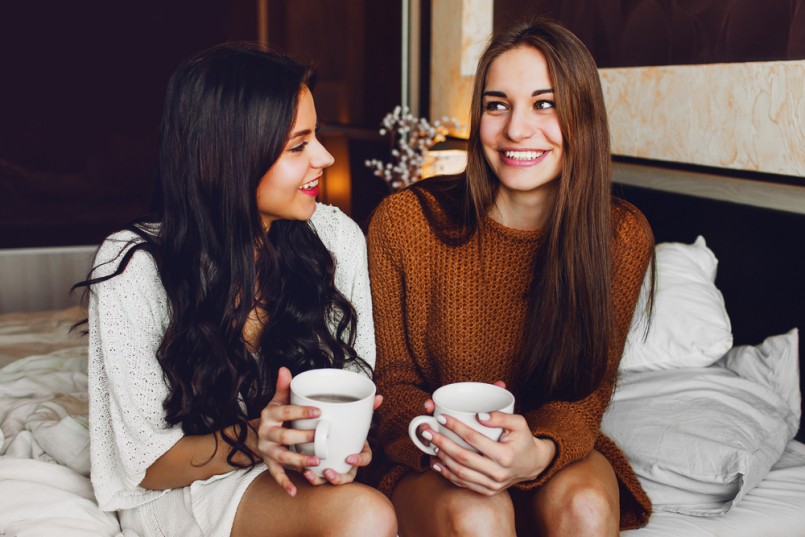Close up lifestyle portrait of two best friends talking and laughing sitting on bed at home. Enjoying sunny morning