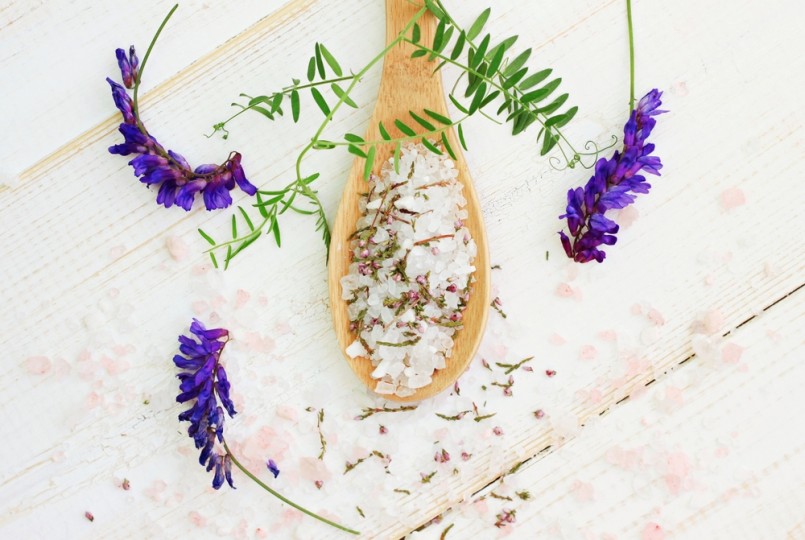 Pink and white bath epsom salt mixture in wooden spoon, fresh and dried aroma herbs. Wooden surface, top view destressing mineral spa background