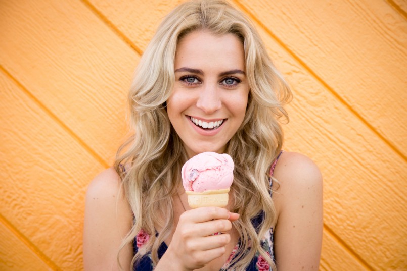 A cute twenty something woman has fun while eating her strawberry ice cream cone while standing in front of an wooden bright orange wall