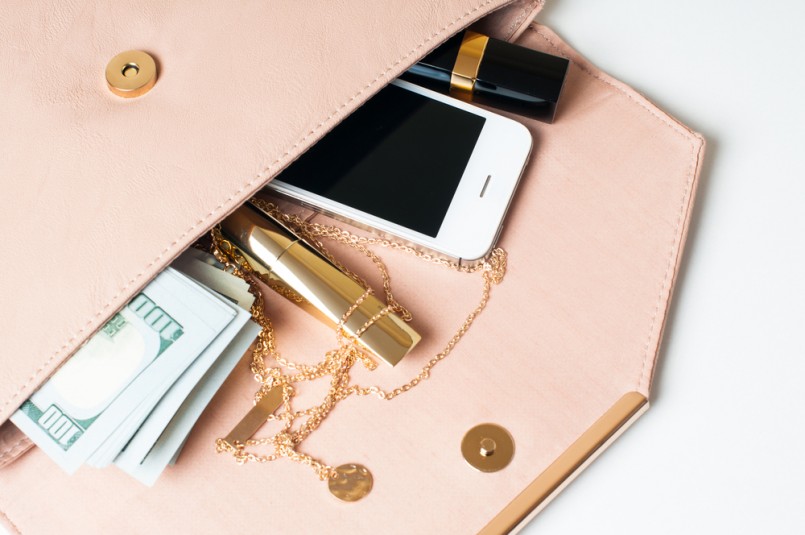 Cosmetics, jewelry, money and smartphone in an open beige woman's clutch handbag on a white background