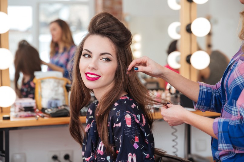 Female hairdresser standing and making hairstyle to cute lovely young woman in beauty salon