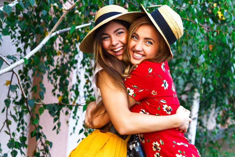 Two happy pretty young sisters, hugs smiling laughing and having funny crazy time together, bearing stylish retro vintage feminine clothes and hats. Outdoors