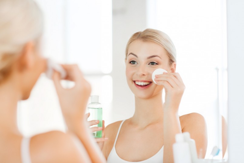 beauty, skin care and people concept - smiling young woman applying lotion to cotton disc for washing her face at bathroom