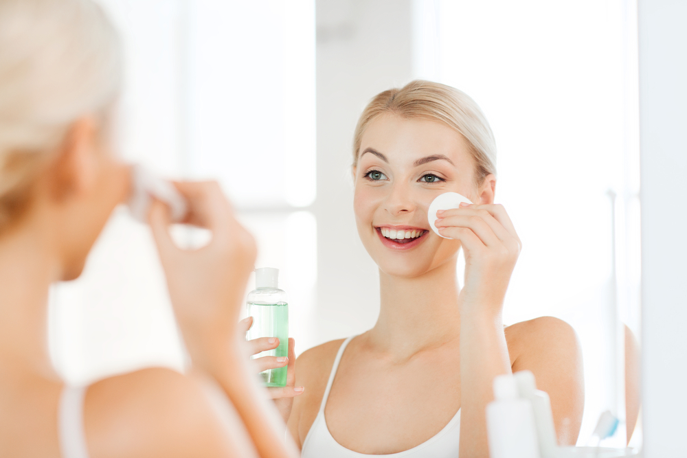 beauty, skin care and people concept - smiling young woman applying lotion to cotton disc for washing her face at bathroom