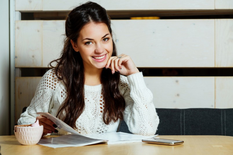 Smiling brunette woman, with curly long hair, wearing in white sweater, sitting at the table and reading a magazine with cup of coffee and smart phone, in cafe with wooden walls, waist up
