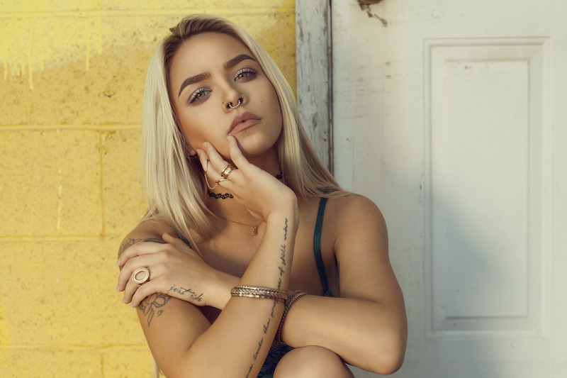 beautiful tan woman with blonde hair and tattoos on skin sitting smoldering yellow wall