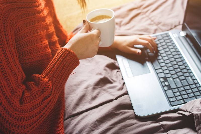 Young brunette woman is lying on her bad in the morning with laptop and a cup of coffee.