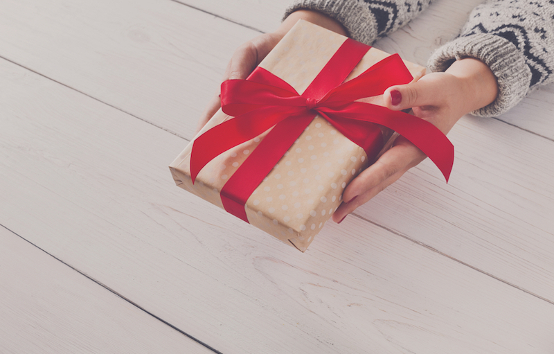 Woman's hands give wrapped christmas or other holiday handmade present in paper with red ribbon. Present box, decoration of gift on white wooden table with copy space